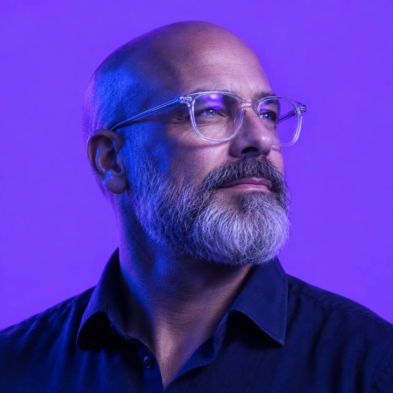 A bald man with a grey beard wearing clear-frame glasses with violet reflections