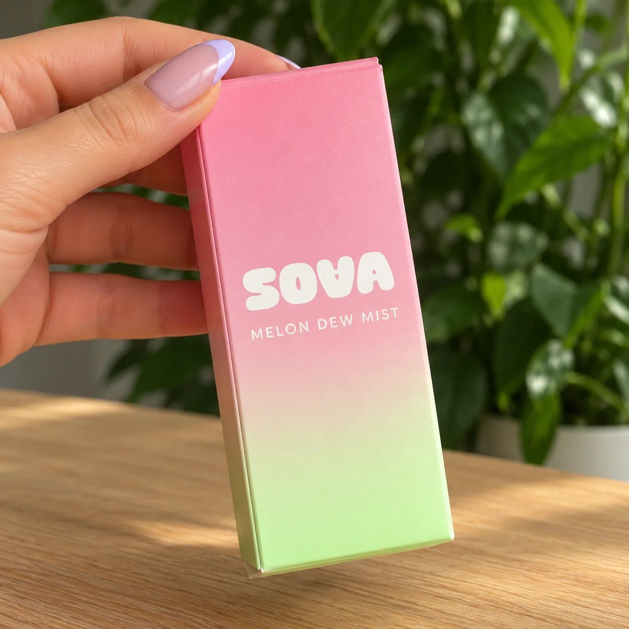 A manicured hand holding a pink-to-lime SOVA Melon Dew Mist carton above a wooden tabletop.