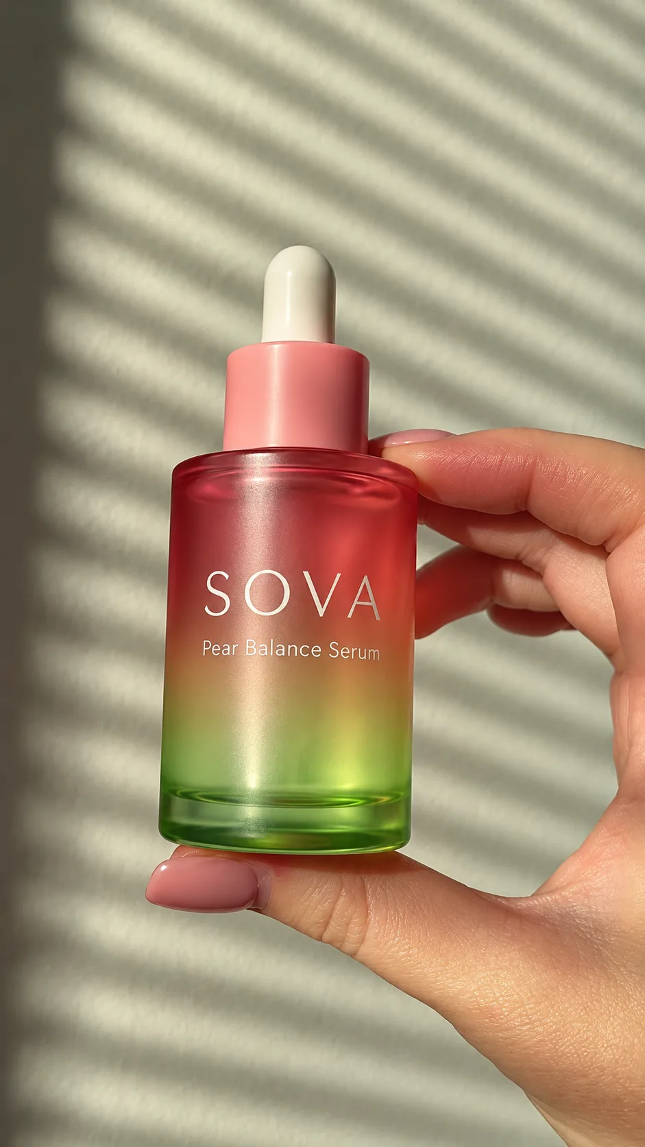 Hand holding a pink-to-green SOVA serum bottle in striped sunlight.